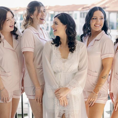 A group of bridesmaids and a bride in matching pink robes laugh together outdoors in sunny wedding-day joy.