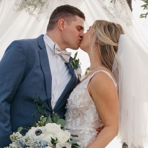 A bride and groom kiss under a white tent, dressed in wedding attire, holding flowers.