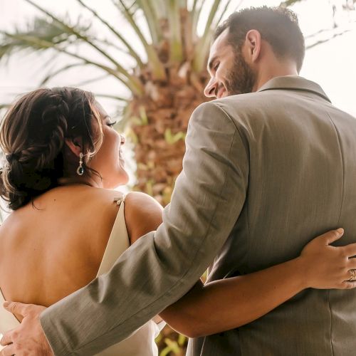 A man in a light suit embraces a woman in a white dress outdoors among palm trees at golden hour.