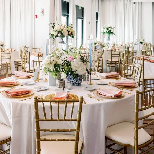 A bright wedding reception setup: round tables with gold chiavari chairs, folded napkins, and white-pink floral centerpieces.