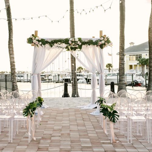 A waterfront wedding setup with a floral arch, white chairs, string lights, and palm trees behind a dock.