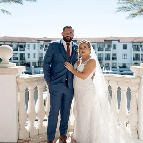 A bride and groom pose smiling on a sunny hotel balcony, dressed in wedding attire against palm trees and a white building.