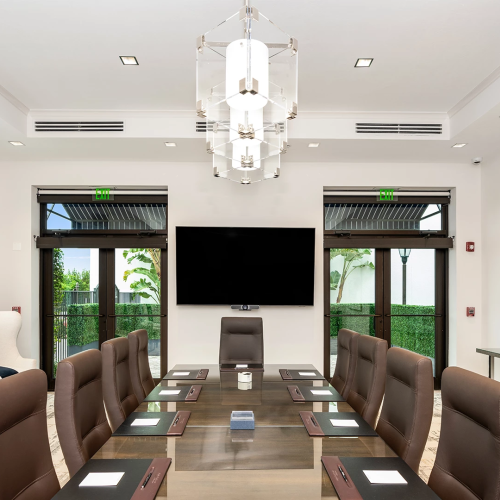 A modern conference room with a long table, brown leather chairs, glass doors to greenery, a wall-mounted TV, chandeliers, and abstract art on the wall.