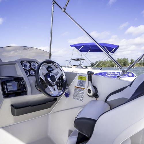 A sunny boat cockpit with a steering wheel and dashboard, blue canopy above, docked on calm water under a clear sky.