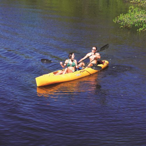 Two people paddle a bright yellow kayak together on a calm river surrounded by green trees.
