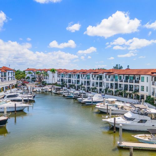 A sunny marina with calm water, lined by white buildings with red-tiled roofs and docked boats on both sides. Ends now.