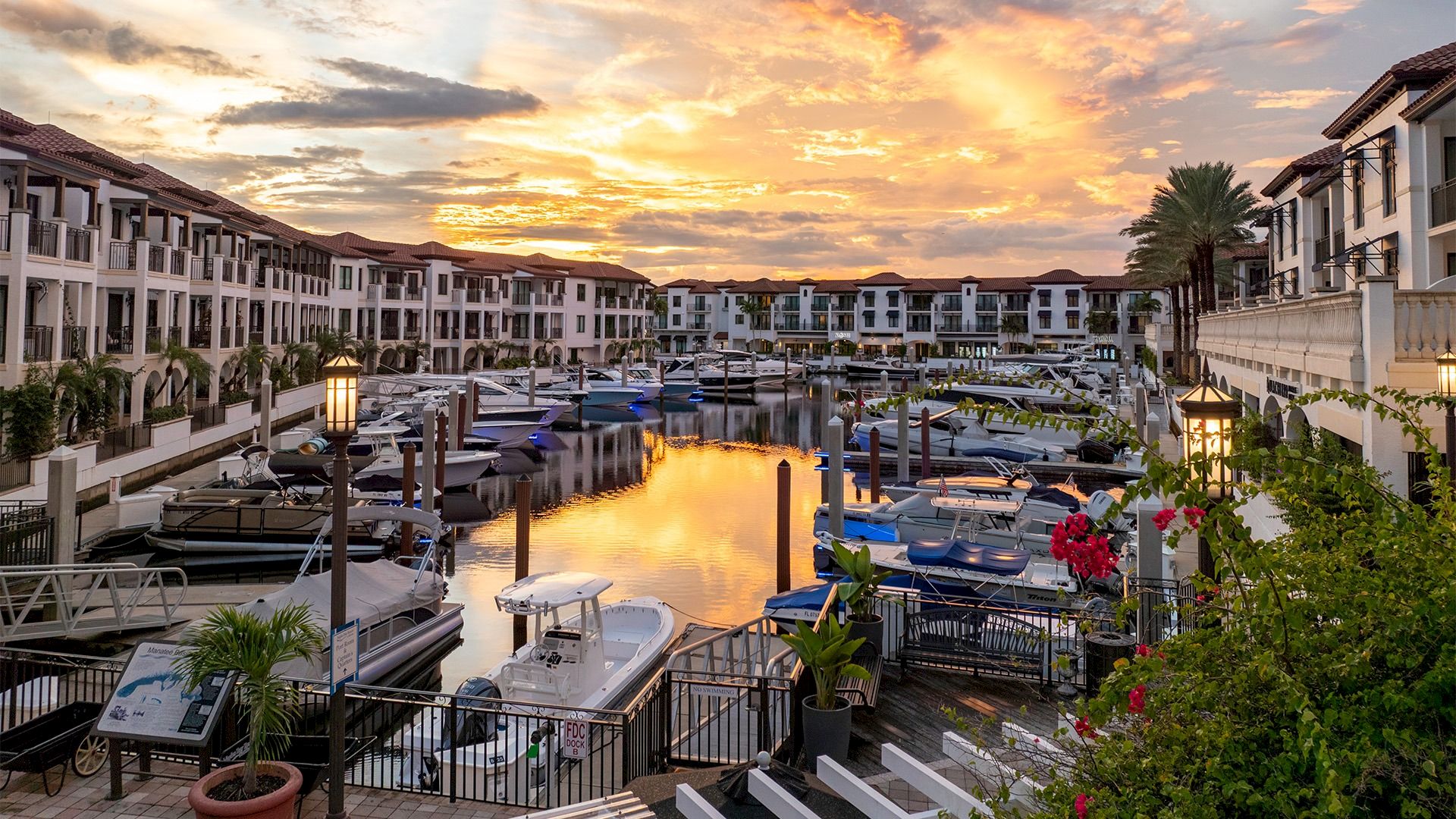 Sunset over a marina with calm water, boats docked, white townhouses, palm trees, and a bridge; warm orange glow reflects off the water.