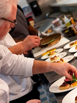 A chef in a white jacket carefully plates gourmet food on a banquet line, adding greens and sauce for guests.