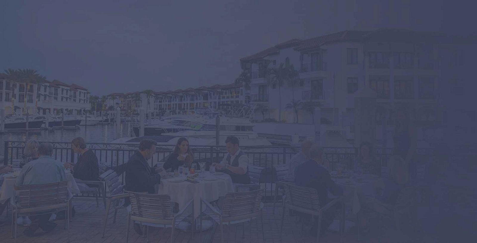 A group of people dining at an outdoor seaside restaurant with tables, chairs, and waterfront buildings in the background.