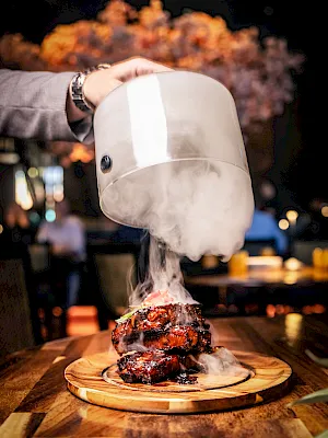 A chef lifts a dome cloche, steaming over a sizzling steak on a wooden board in a smoky restaurant setting.