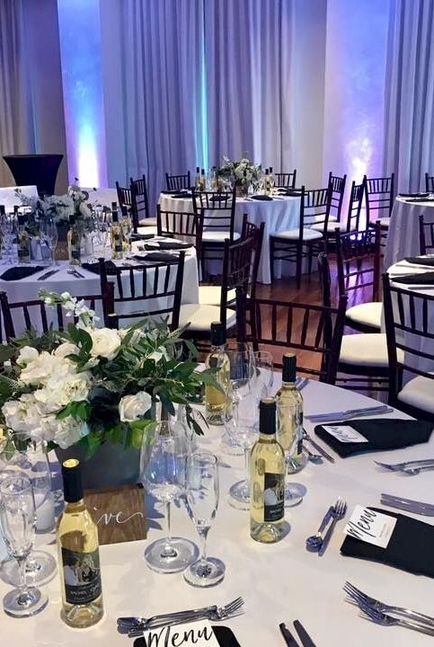 An elegant banquet setup with white tablecloths, black chairs, floral centerpieces, wine bottles, and silver cutlery.