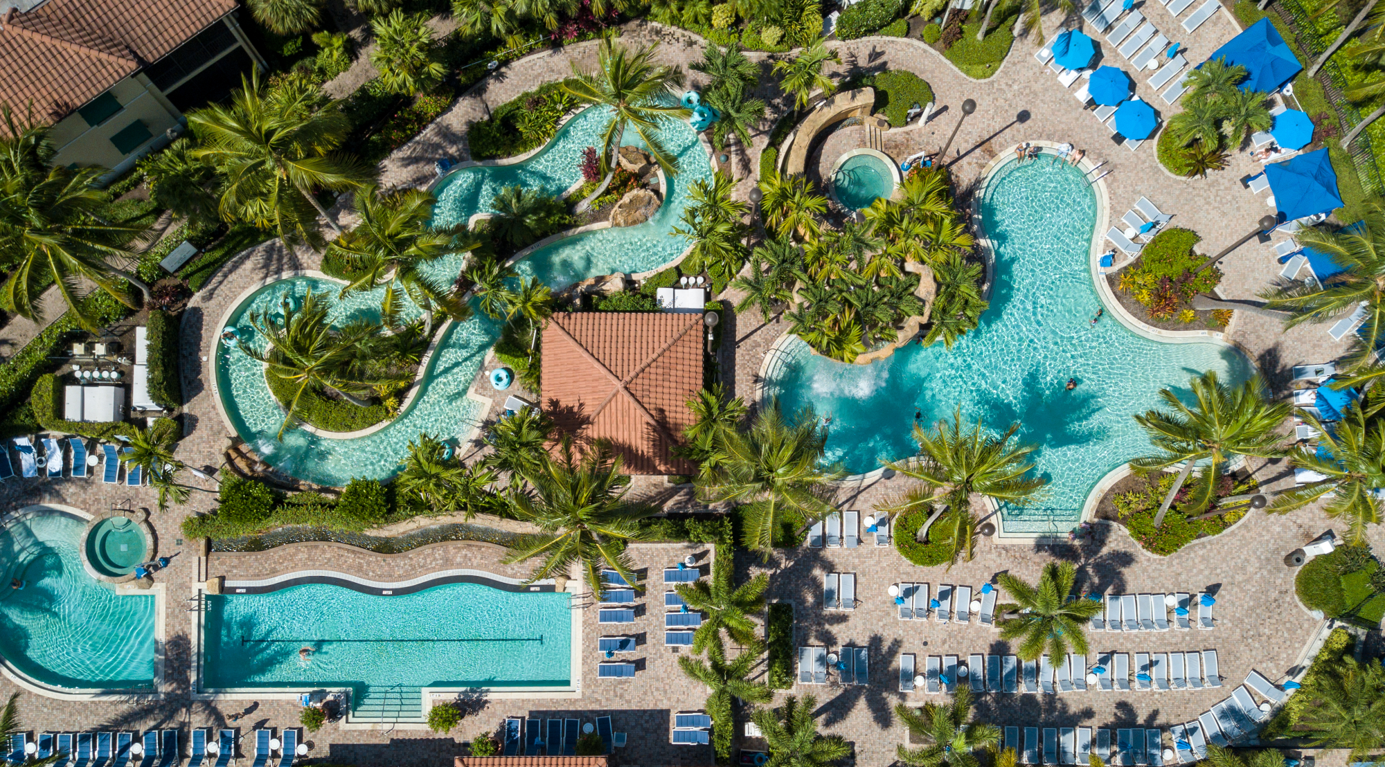 Aerial view of a tropical resort with multiple turquoise pools, palm trees, and rows of blue sunbeds and umbrellas, always.