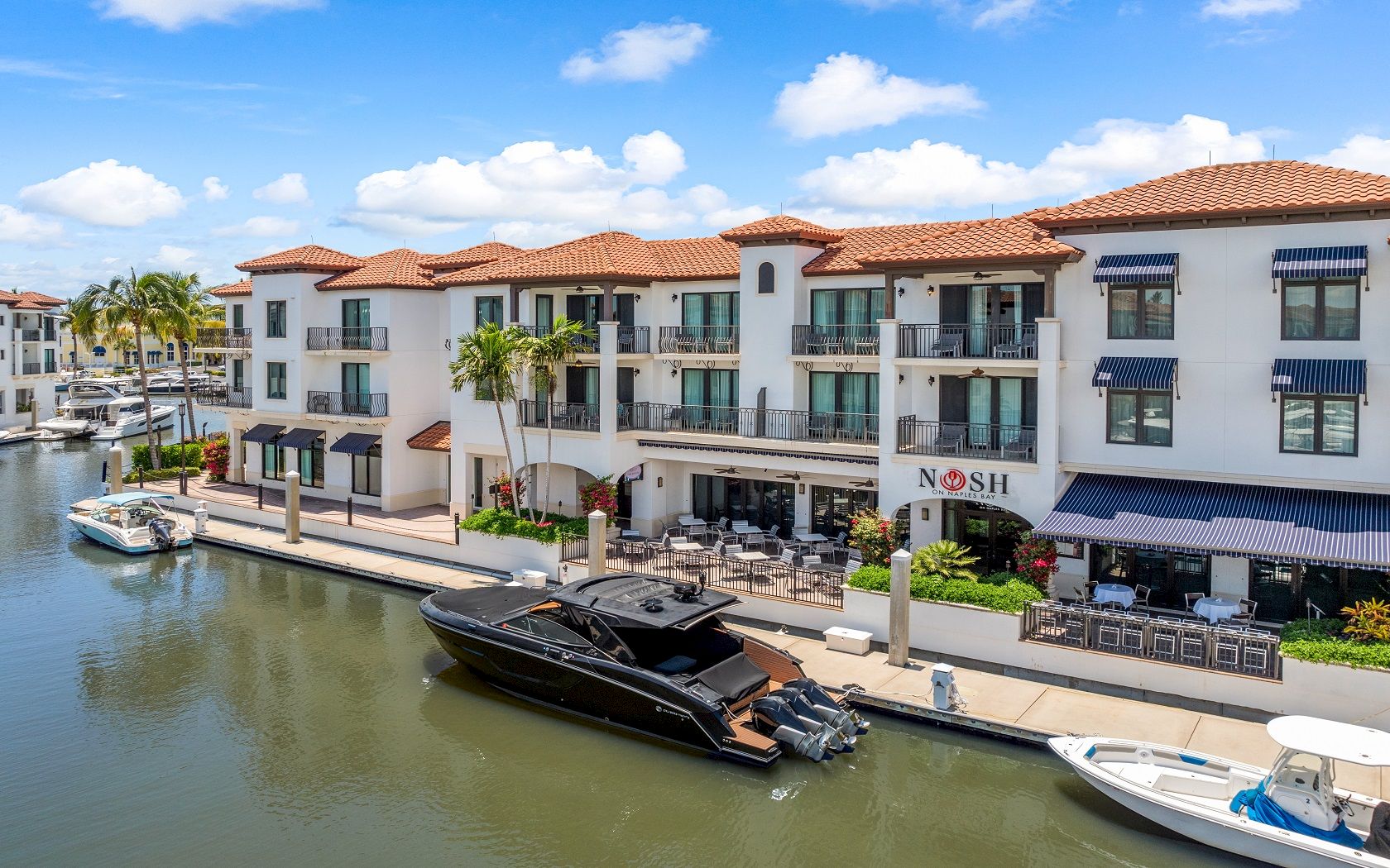 A waterfront hotel complex with terracotta-roof buildings, solar awnings, manicured gardens, and yachts docked along the canal.