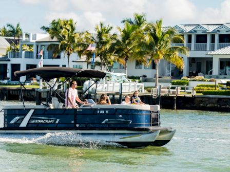 A pontoon boat cruises on calm water with a few passengers near palm trees and waterfront homes under a blue sky.