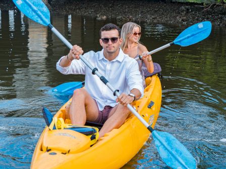 Couple in sunglasses paddles a yellow kayak with blue paddles under a bridge on a calm river.