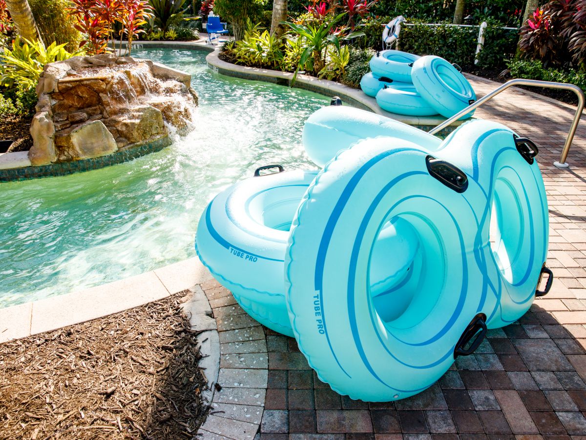 A bright blue inflatable pool tube sits by a garden fountain-style pool, surrounded by lush tropical plants and palm trees.