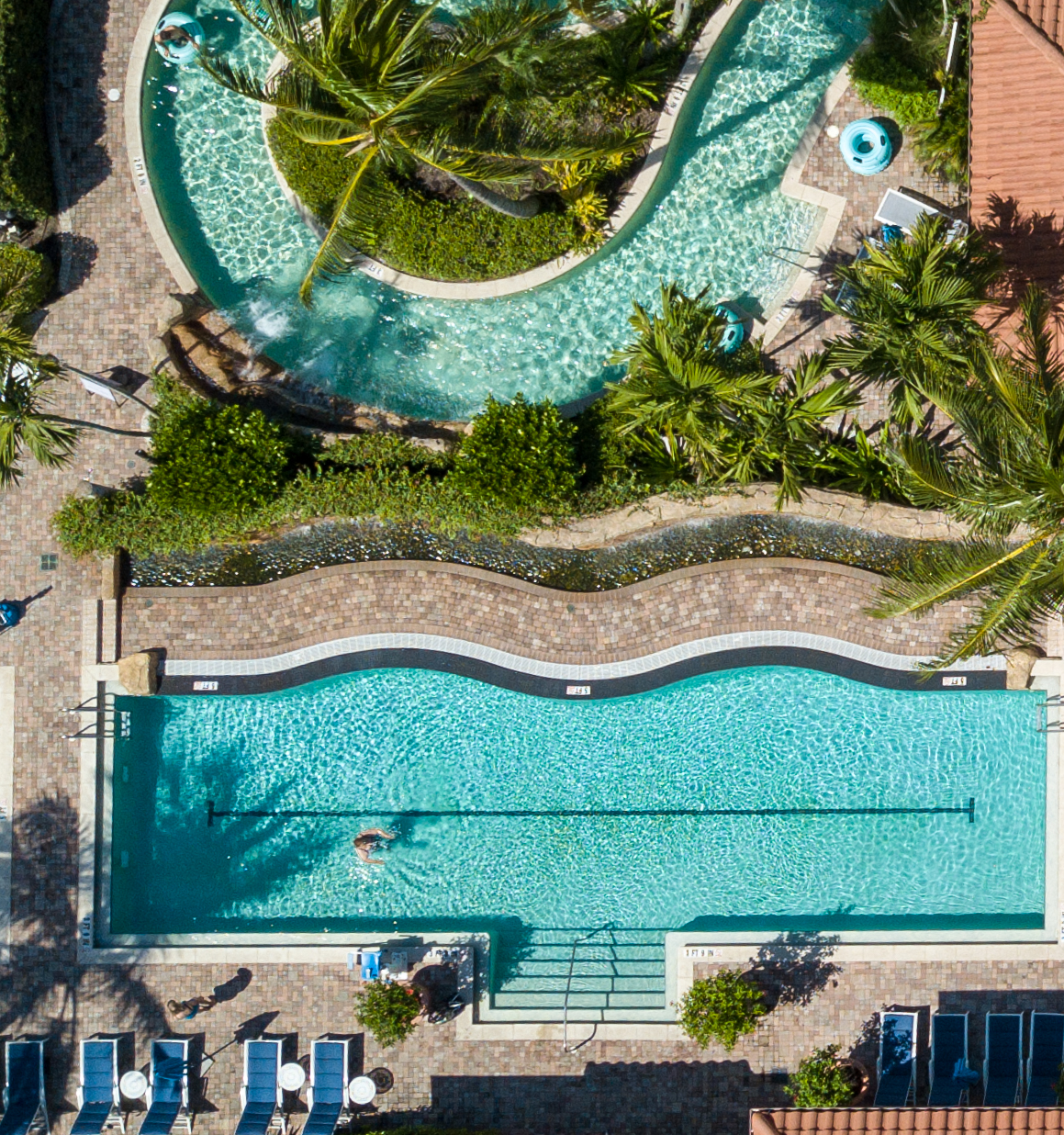 Aerial view of a tropical resort with two swimming pools, a circular whirlpool, palm trees, and sun loungers.