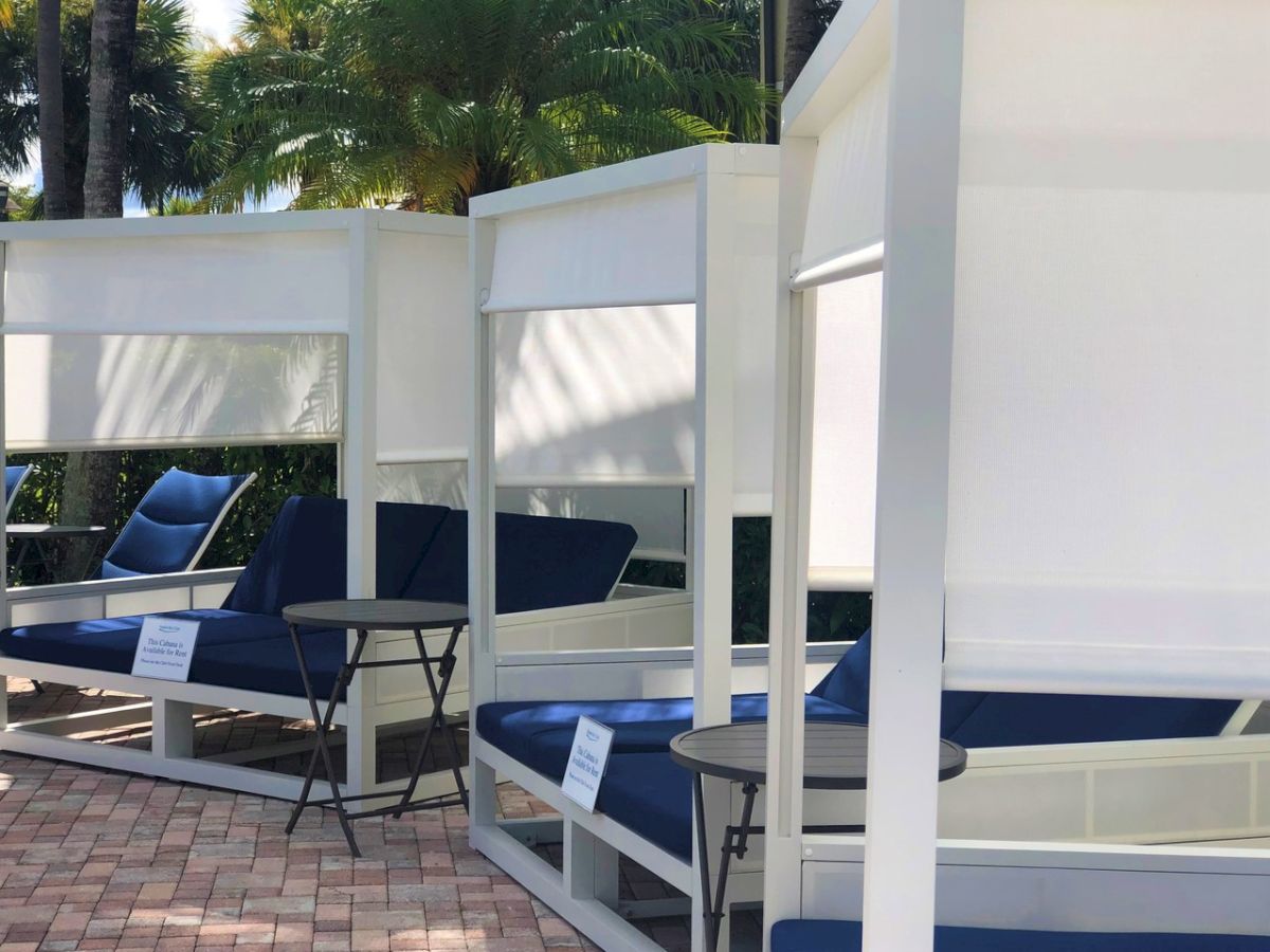 A sunny poolside lounge with white cabanas, navy cushioned seating, round tables, and palm trees in the background.