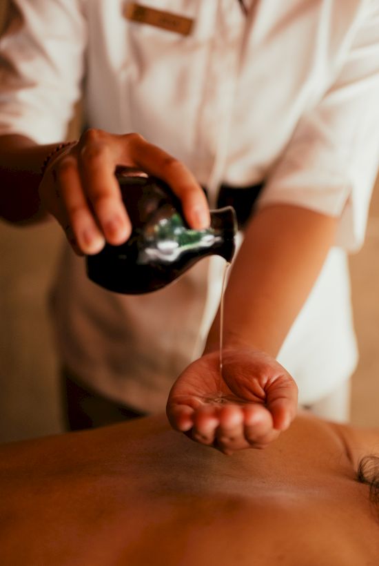 A therapist performs a massage, applying oil to a person&rsquo;s bare back with a handheld tool.