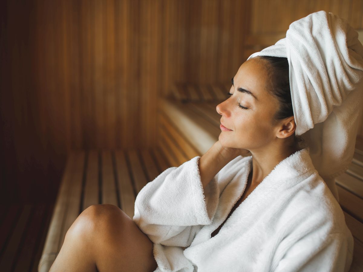 A woman relaxing in a warm sauna, wrapped in a white robe with a towel on her head, eyes closed.