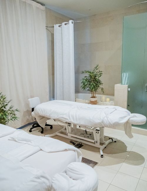 A bright spa treatment room with curtained massage beds, a potted plant, and a glass sink area, lit overhead.