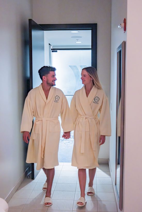 A man and woman in matching spa robes talk in a hallway doorway, facing each other before a bright room.