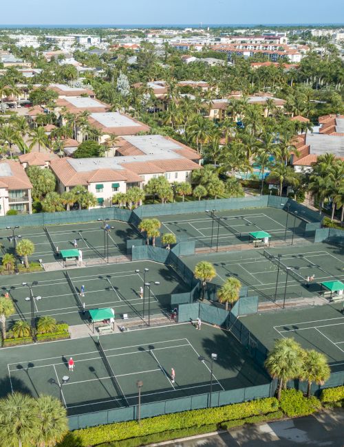 Aerial view of a tropical seaside resort with palm trees, red-tiled villas, and multiple fenced tennis courts, plus the ocean in back.