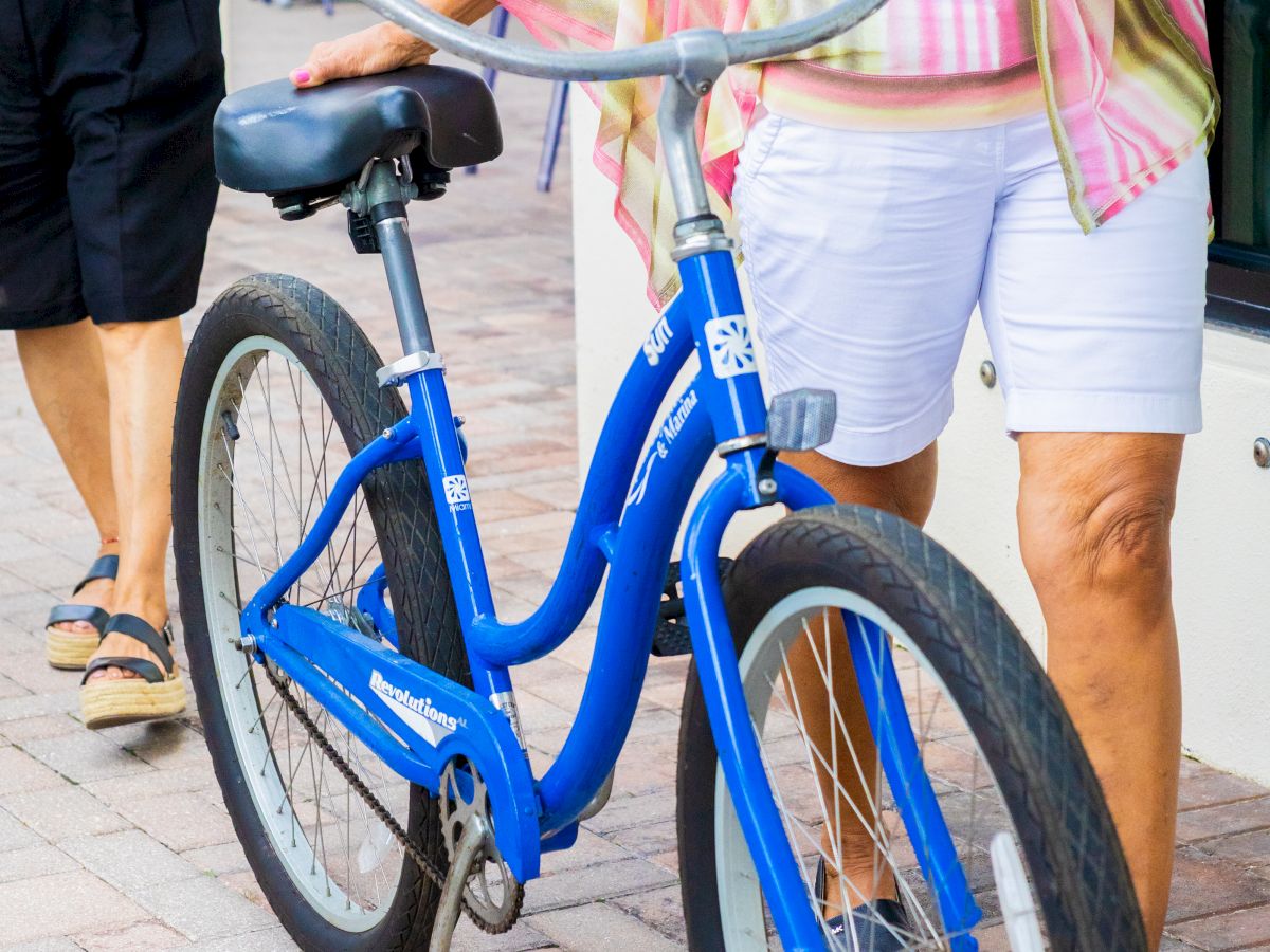 A person adjusts a blue rental bicycle while another waits nearby on a sunny patio.