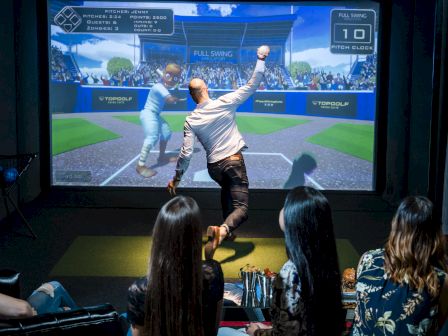 A person swings a golf club at a Topgolf-style simulator, with a large projected scoreboard and crowd background on-screen.