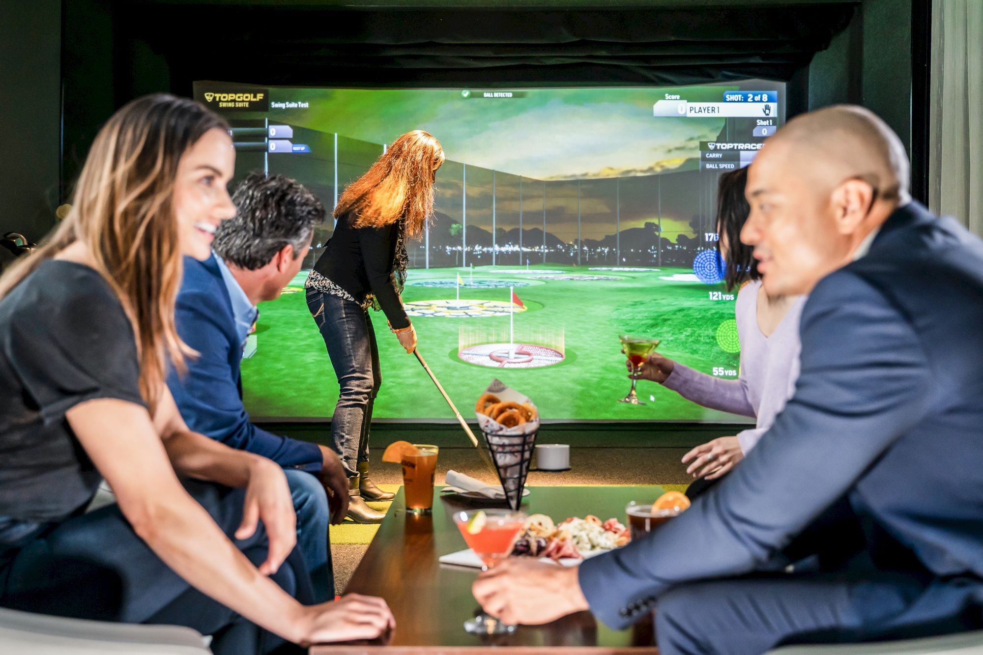 People sit at a table watching a golf simulator screen as a player aims a club at the virtual green.