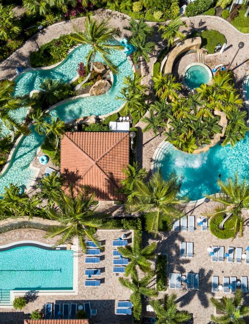 Aerial view of a tropical resort: multiple turquoise pools, winding waterslides, palm trees, and blue cabanas with loungers.