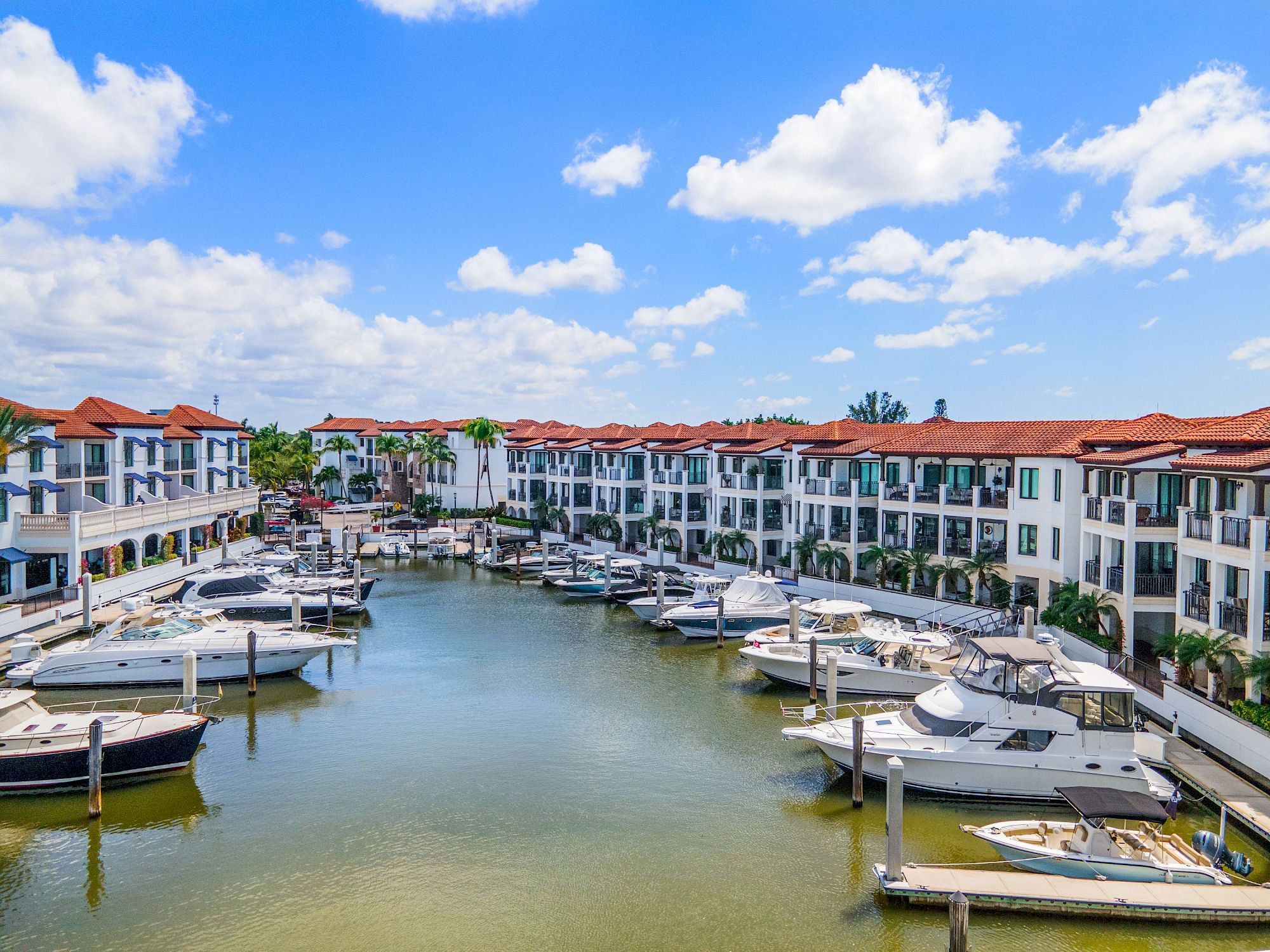 A sunny canal marina with docked yachts and white resort buildings beneath a bright blue sky and fluffy clouds.
