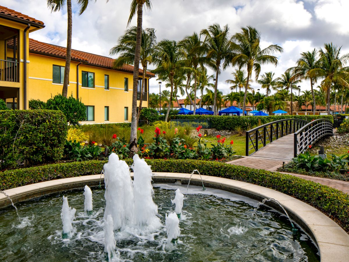 A tropical resort courtyard with a bubbling fountain, palm trees, colorful flowers, and yellow buildings with blue shade tents.