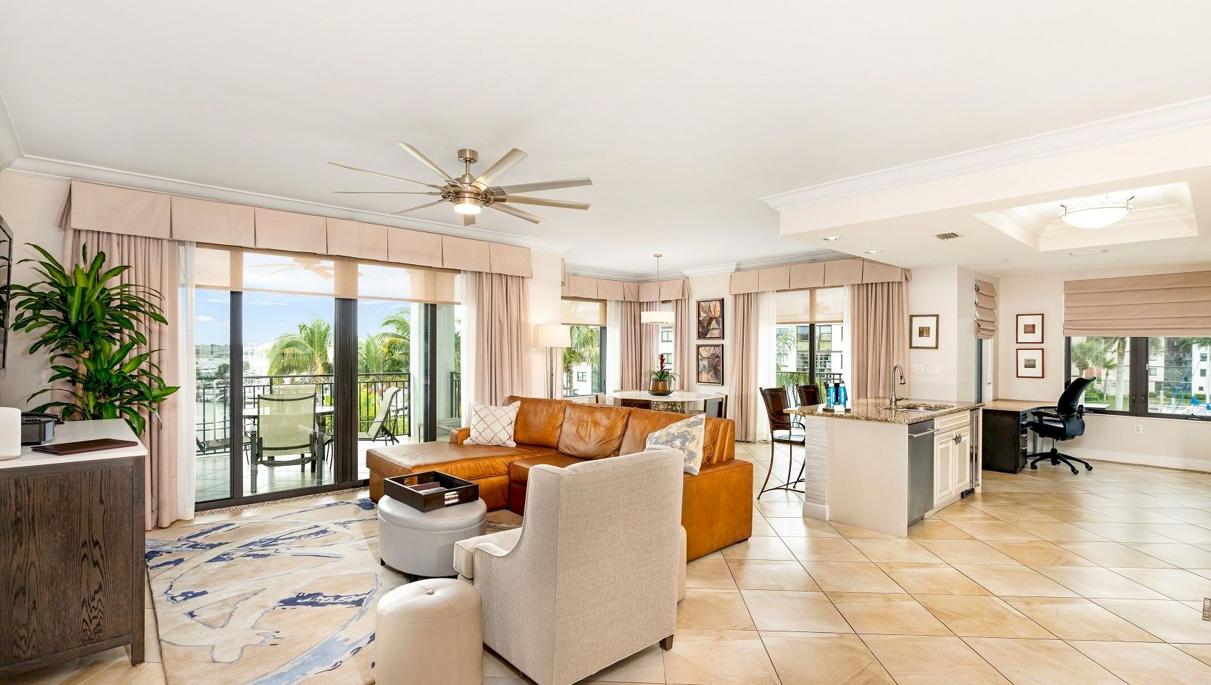 Bright open-plan living room with sofas, ceiling fan, large windows, and a kitchen island with chairs.
