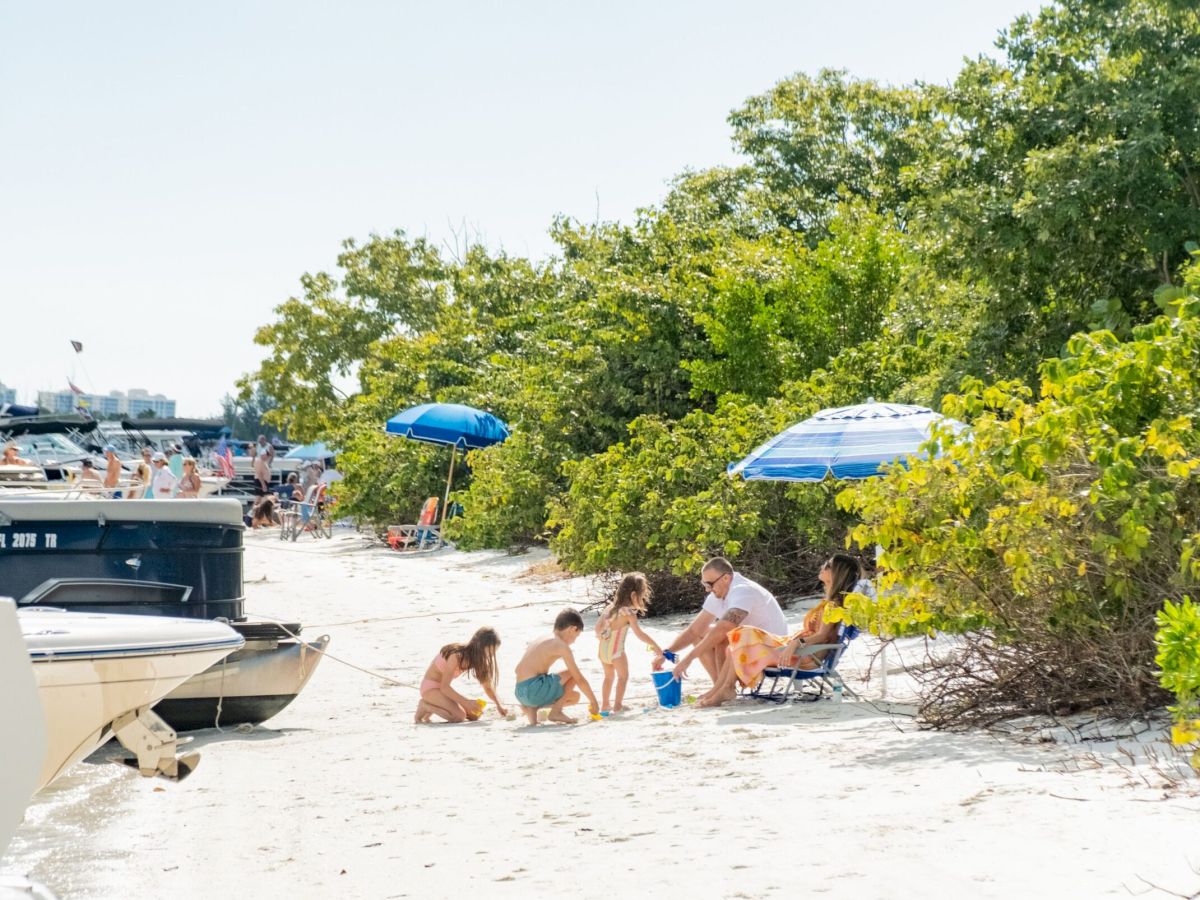 A sunny beach scene with people playing in the sand, boats at the shore, and blue umbrellas beside green bushes.