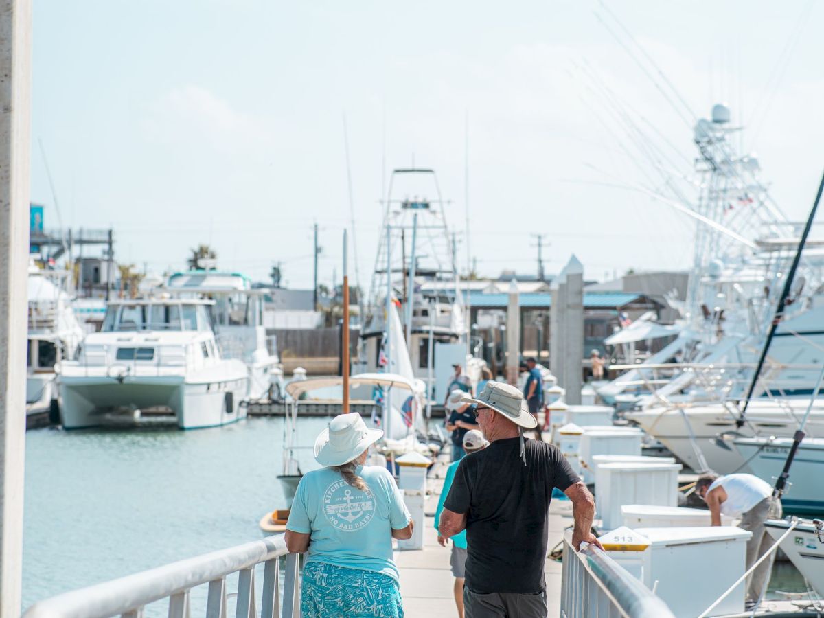 A marina pier with two people walking past parked boats and yachts on calm blue water under a clear sky.