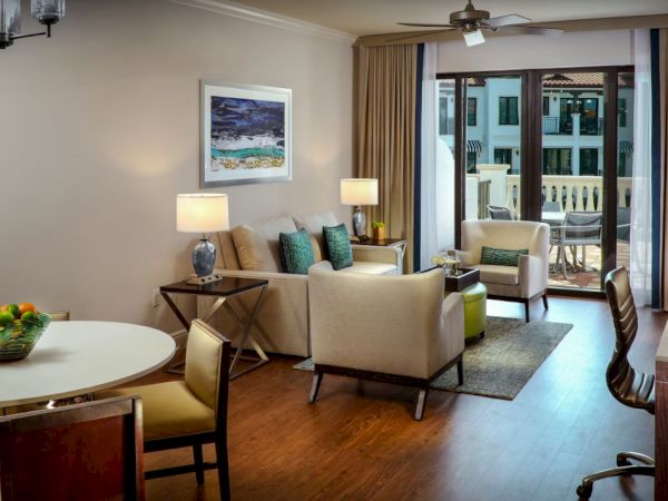 Bright modern living room with beige sofas, lamps, a dining table, and patio doors showing an outdoor balcony.
