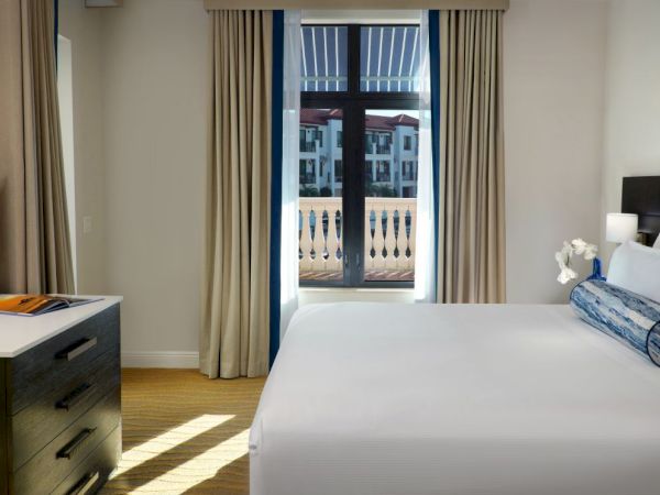 A bright hotel bedroom with a neatly made bed, bedside dresser, and curtains opening to a view outside.