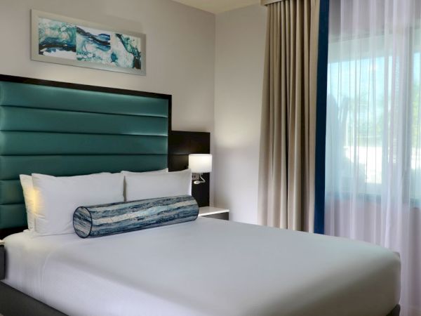 A modern hotel bedroom with a neatly made bed, bedside tables, and bright window curtains letting in daylight.