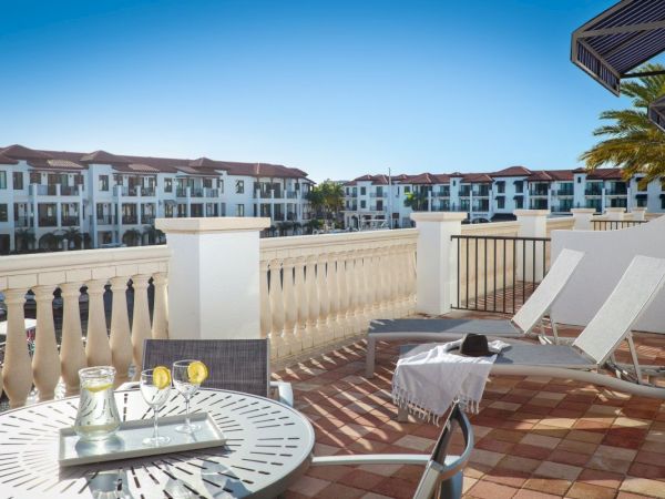 A resort balcony with a round table, drinks, sun loungers, and a view of white townhouses under a clear blue sky.