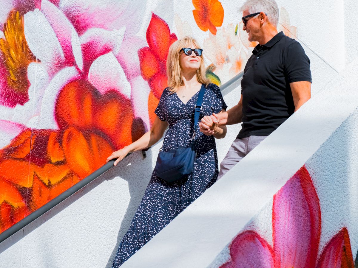 A woman and a man pose on outdoor steps beside a colorful graffiti mural, with the man in black and the woman in sunglasses.