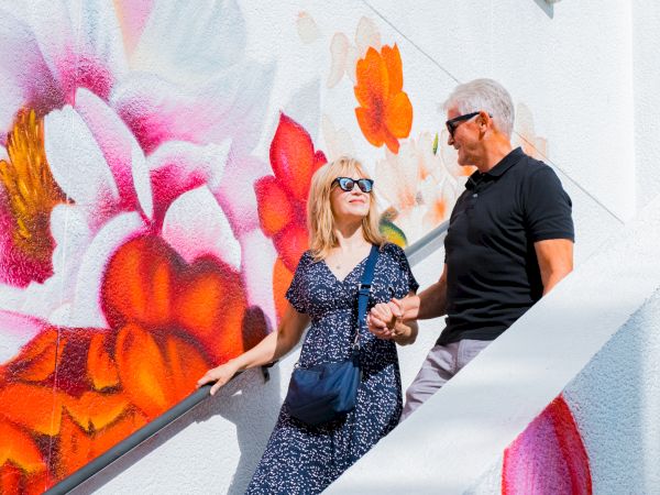 A man and woman walk hand-in-hand by a white wall covered in colorful graffiti, smiling as they pose by the mural-filled stairs.