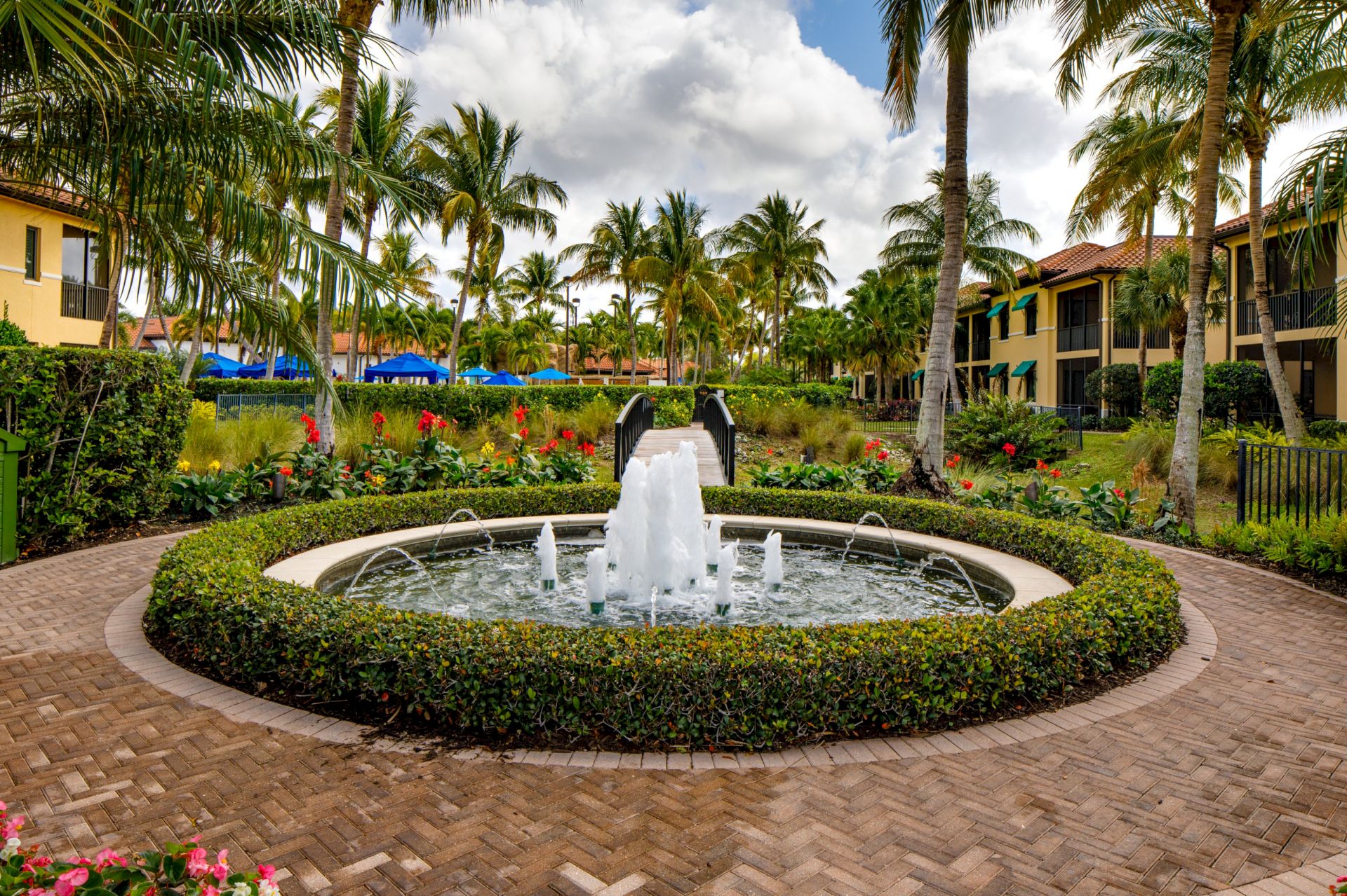 Tropical resort courtyard with palm trees, flower beds, and a circular fountain splashing under cloudy skies.