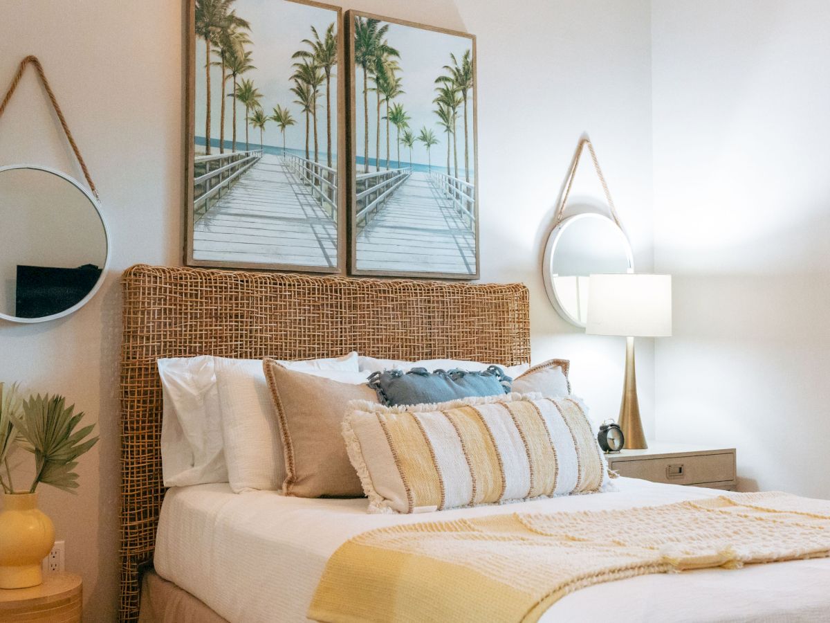A cozy bedroom with a rattan headboard, layered pillows and throw, bedside lamps, a circular mirror, and palm-print wall art.